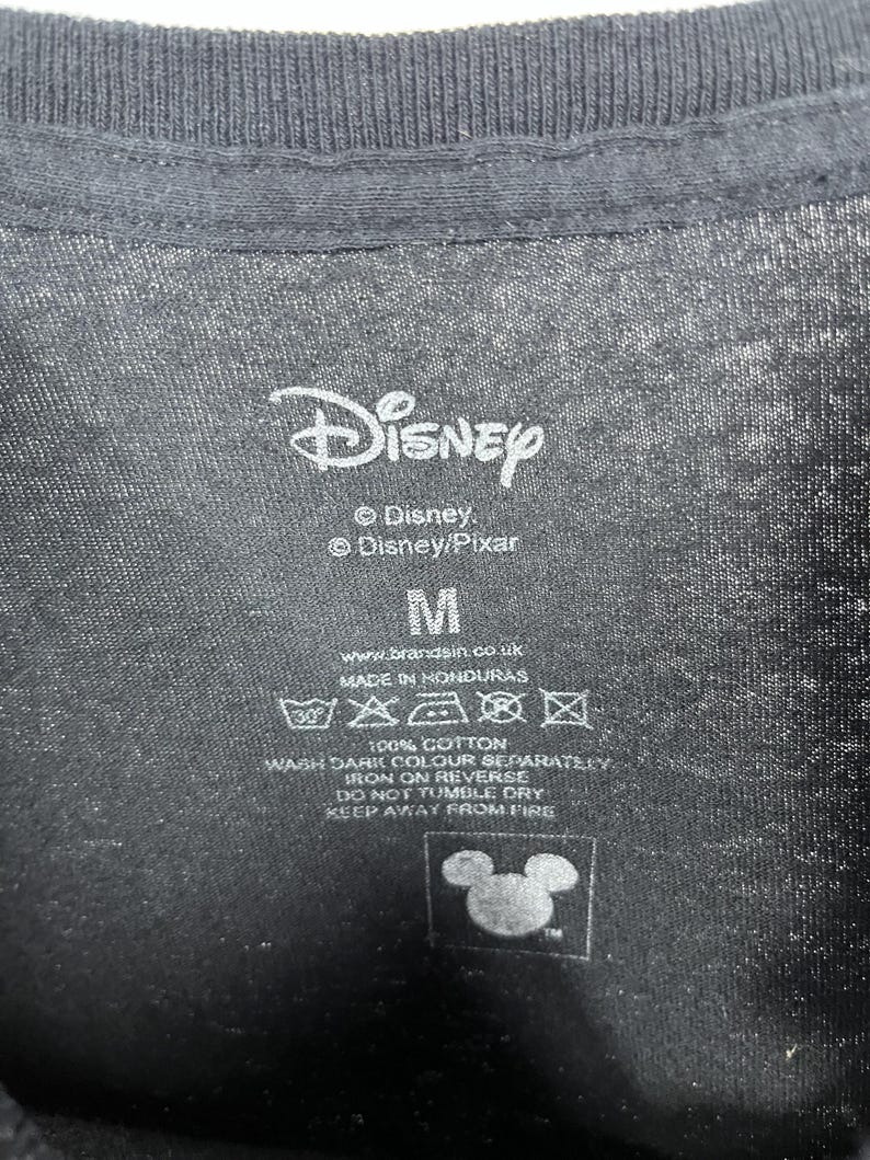 Peut inclure: Gros plan sur une &eacute;tiquette de t-shirt noire avec le logo Disney et les textes "Disney" et "Disney/Pixar". L'&eacute;tiquette indique &eacute;galement la taille "M", des instructions de lavage et le texte "100% Coton".