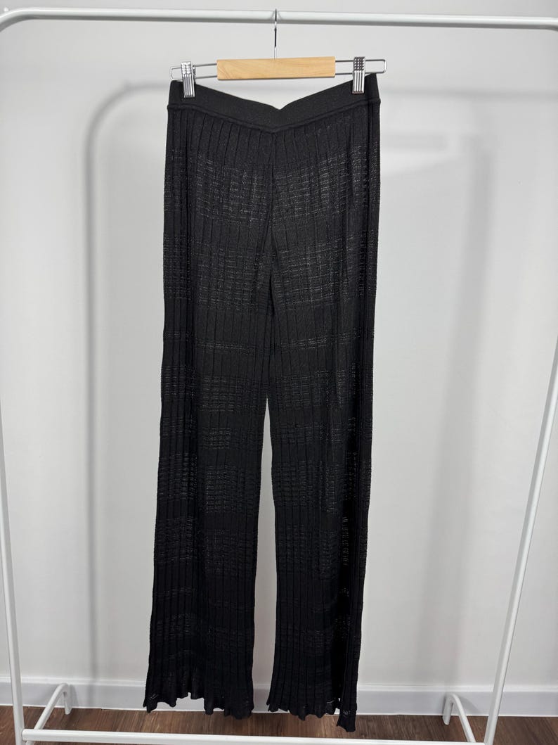 May include: Black, wide-leg pants with a ribbed texture, hanging on a hanger. The pants have a high waist and a slight flare at the hem. The fabric appears to be lightweight and semi-sheer.