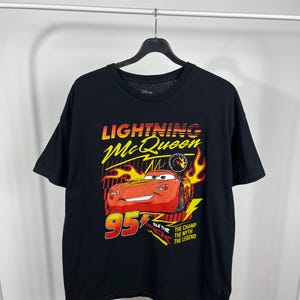 Peut inclure: T-shirt noir avec un motif de Flash McQueen du film Cars de Disney. Le graphisme comprend le texte "Lightning McQueen" en jaune et orange, le num&eacute;ro "95" en rouge et les phrases "The Champ, The Myth, The Legend."