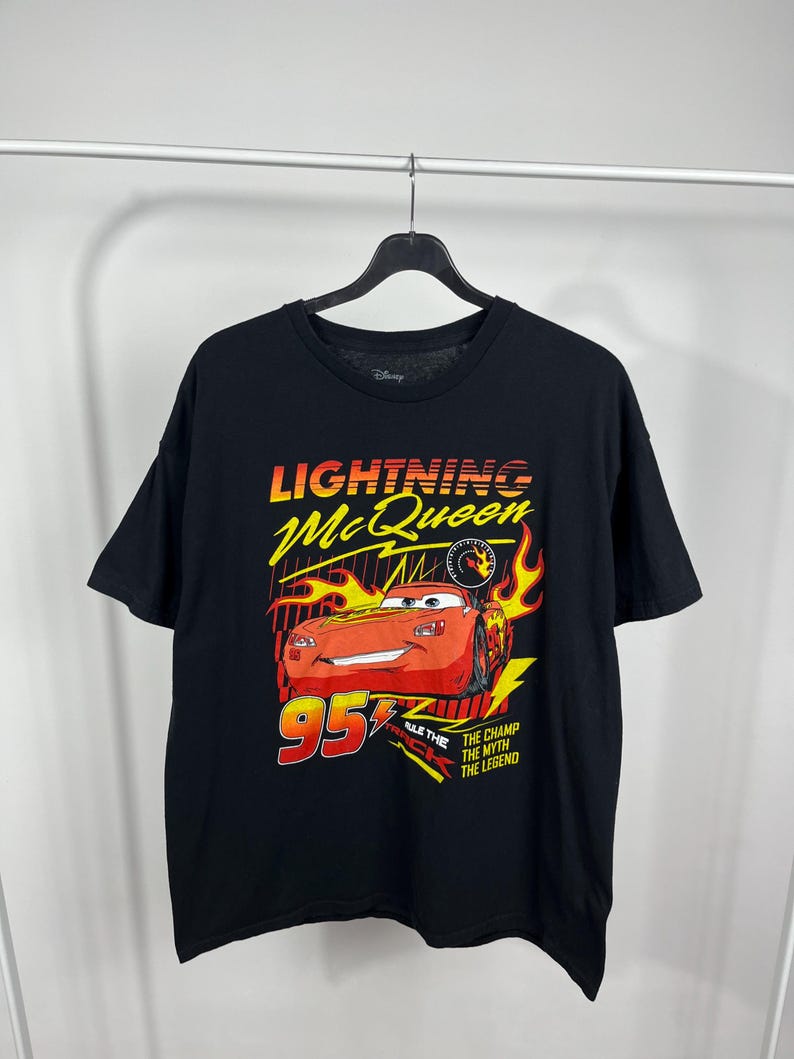 Peut inclure: T-shirt noir avec un motif de Flash McQueen du film Cars de Disney. Le graphisme comprend le texte "Lightning McQueen" en jaune et orange, le num&eacute;ro "95" en rouge et les phrases "The Champ, The Myth, The Legend."