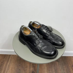 Peut inclure: Une paire de chaussures de ville en cuir noir avec des lacets, présentée sur une petite table ronde grise. Les chaussures ont un design classique avec un bout arrondi et un petit talon. Le fond est un mur blanc uni et un sol en bois.