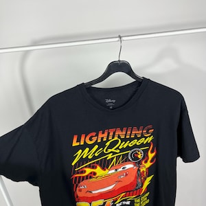 Peut inclure: T-shirt noir avec un motif de Flash McQueen du film Cars de Disney. Le graphisme comprend le texte "Lightning McQueen" en jaune et rouge, le num&eacute;ro "95" et des flammes. Le t-shirt est suspendu &agrave; un cintre noir.