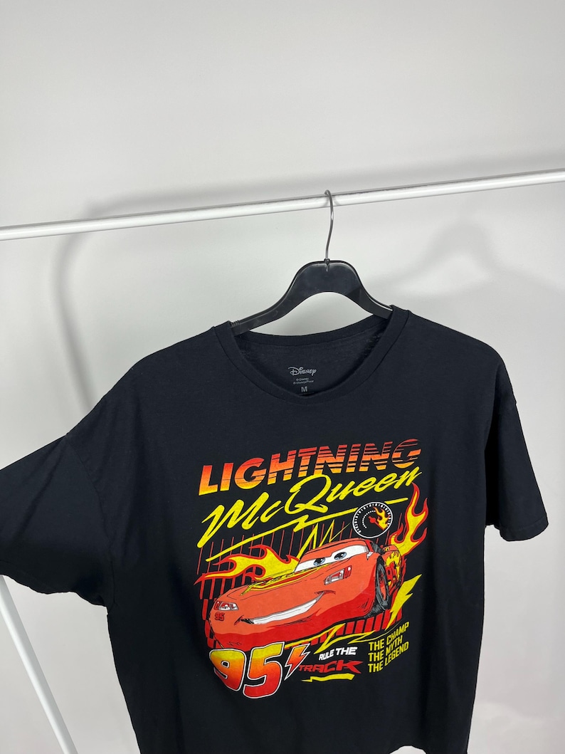 Peut inclure: T-shirt noir avec un motif de Flash McQueen du film Cars de Disney. Le graphisme comprend le texte "Lightning McQueen" en jaune et rouge, le num&eacute;ro "95" et des flammes. Le t-shirt est suspendu &agrave; un cintre noir.