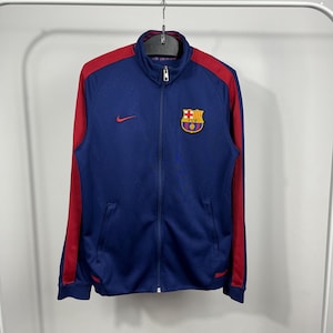 May include: A navy blue zip-up track jacket with red shoulder and sleeve accents. Features the FC Barcelona team crest on the chest and a small red Nike logo. The jacket has a stand-up collar and two front pockets.