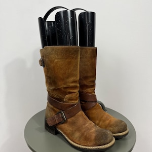 Puede incluir: Un par de botas de ante marrón con correas de cuero y hebillas. Las botas tienen un aspecto desgastado y están sobre una pequeña mesa redonda gris. Las botas son altas, llegando a la pantorrilla, unos 30 cm.