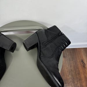 Pu&ograve; includere: Stivaletti in pelle nera con tacco a blocco e zip laterale. Gli stivali hanno la punta arrotondata e sono su una superficie grigia. L'altezza del tacco &egrave; di circa 7,6 cm.