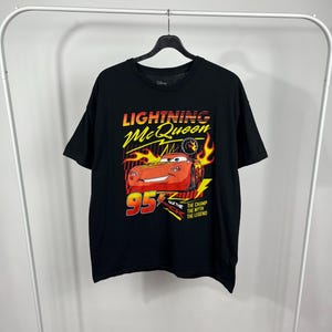 Peut inclure: T-shirt noir avec un motif de Flash McQueen du film Disney Cars. Le design comprend McQueen, le num&eacute;ro 95 et le texte "The Champ, The Myth, The Legend". Les couleurs sont rouge, jaune et blanc.