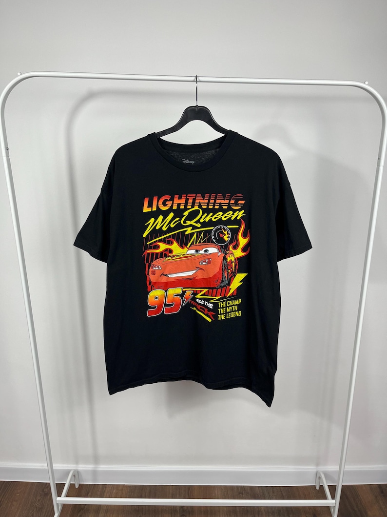 Peut inclure: T-shirt noir avec un motif de Flash McQueen du film Disney Cars. Le design comprend McQueen, le num&eacute;ro 95 et le texte "The Champ, The Myth, The Legend". Les couleurs sont rouge, jaune et blanc.