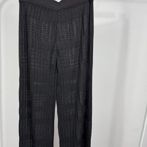 May include: Black, wide-leg pants with a textured, ribbed design hanging on a clothing rack. The pants have a high waist and a subtle sheen, suggesting a knit or woven fabric. The fabric appears to have a grid-like pattern.