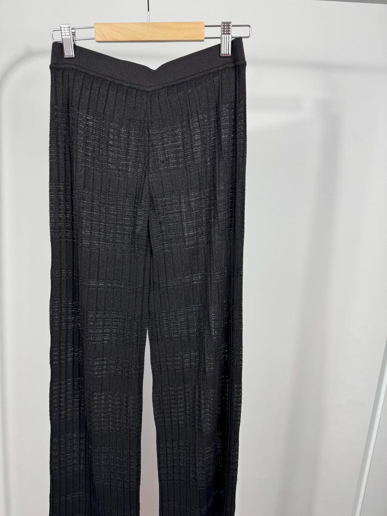 May include: Black, wide-leg pants with a textured, ribbed design hanging on a clothing rack. The pants have a high waist and a subtle sheen, suggesting a knit or woven fabric. The fabric appears to have a grid-like pattern.