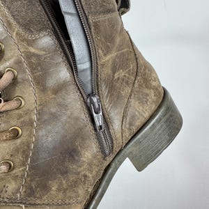 K&ouml;nnte beinhalten: Ein kn&ouml;chelhoher Stiefel aus braunem, abgenutztem Leder mit seitlichem Rei&szlig;verschluss, messingfarbenen Metall&ouml;sen und dicken braunen Schn&uuml;rsenkeln. Der Stiefel hat einen niedrigen Blockabsatz und sichtbare N&auml;hte, die eine abgenutzte Textur aufweisen.