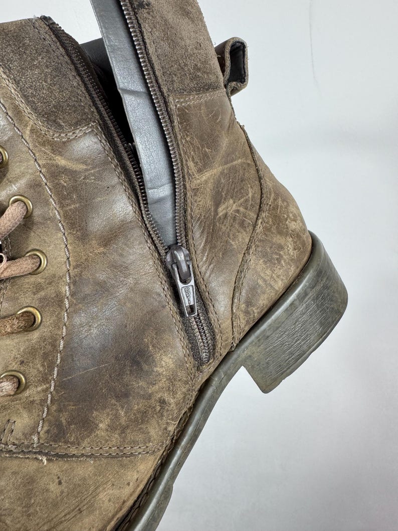 K&ouml;nnte beinhalten: Ein kn&ouml;chelhoher Stiefel aus braunem, abgenutztem Leder mit seitlichem Rei&szlig;verschluss, messingfarbenen Metall&ouml;sen und dicken braunen Schn&uuml;rsenkeln. Der Stiefel hat einen niedrigen Blockabsatz und sichtbare N&auml;hte, die eine abgenutzte Textur aufweisen.