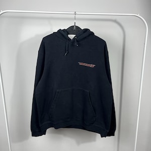 Puede incluir: Sudadera negra con bolsillo delantero y capucha. La sudadera tiene el texto "TEKKEN 5" en rojo en el pecho. La prenda está colgada en una percha blanca.