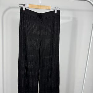 May include: Black, wide-leg pants with a textured, ribbed design. The pants are hanging on a wooden hanger against a white background. The fabric appears to have a subtle sheen.