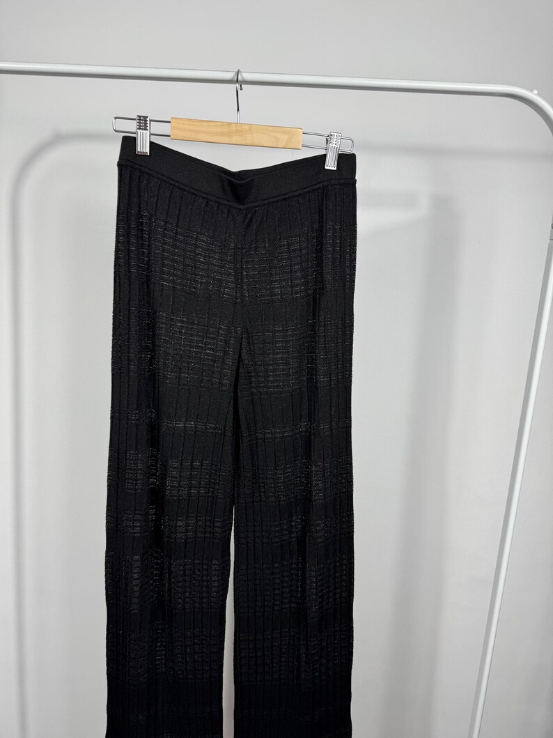 May include: Black, wide-leg pants with a textured, ribbed design. The pants are hanging on a wooden hanger against a white background. The fabric appears to have a subtle sheen.