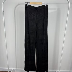 May include: Black knit pants hanging on a white clothing rack. The pants have a wide leg design and a textured, open weave pattern. The waistband is solid black. The pants are made of a lightweight material.