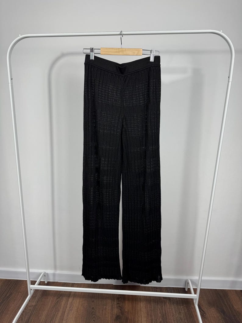May include: Black knit pants hanging on a white clothing rack. The pants have a wide leg design and a textured, open weave pattern. The waistband is solid black. The pants are made of a lightweight material.