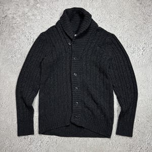 Könnte beinhalten: Eine dunkel anthrazitfarbene Langarm-Strickjacke mit einem dicken, gerippten Schalkragen, einem vertikalen Zopfstrickmuster auf der Vorderseite und den Ärmeln sowie einer Knopfleiste mit fünf passenden dunklen Knöpfen in der Mitte.
