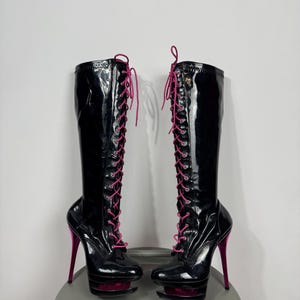 Könnte beinhalten: Ein Paar kniehohe Stiefel aus schwarzem Lackleder mit Schnürung vorne in leuchtendem Pink, einem hohen Stiletto-Absatz in Metallic-Pink und einer Plateausohle mit einer Aussparung, die ein pinkfarbenes Innendetail zeigt.