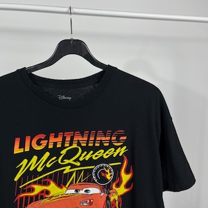 Peut inclure: T-shirt noir avec Lightning McQueen du film Disney Cars. Le graphisme comprend le texte "Lightning McQueen" en jaune et rouge, avec une image de dessin anim&eacute; de la voiture de course et des flammes.