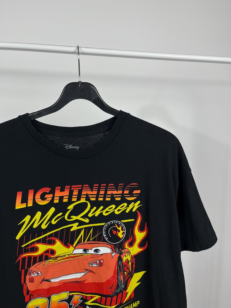 Peut inclure: T-shirt noir avec Lightning McQueen du film Disney Cars. Le graphisme comprend le texte "Lightning McQueen" en jaune et rouge, avec une image de dessin anim&eacute; de la voiture de course et des flammes.