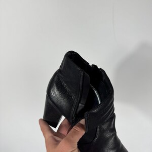 Pu&ograve; includere: Stivaletto alla caviglia in pelle nera con tacco a blocco. Lo stivaletto presenta una cerniera laterale e una tomaia in pelle strutturata. Il tacco &egrave; alto circa 7,6 cm. Lo stivaletto &egrave; tenuto da una mano su sfondo bianco.
