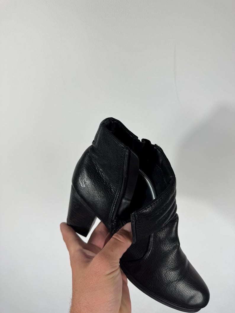 Pu&ograve; includere: Stivaletto alla caviglia in pelle nera con tacco a blocco. Lo stivaletto presenta una cerniera laterale e una tomaia in pelle strutturata. Il tacco &egrave; alto circa 7,6 cm. Lo stivaletto &egrave; tenuto da una mano su sfondo bianco.