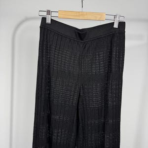 May include: Black, wide-leg pants with a textured, ribbed design. The pants are hanging on a wooden hanger with a silver metal clip. The fabric has a subtle sheen, and the pants appear to be made of a lightweight material.