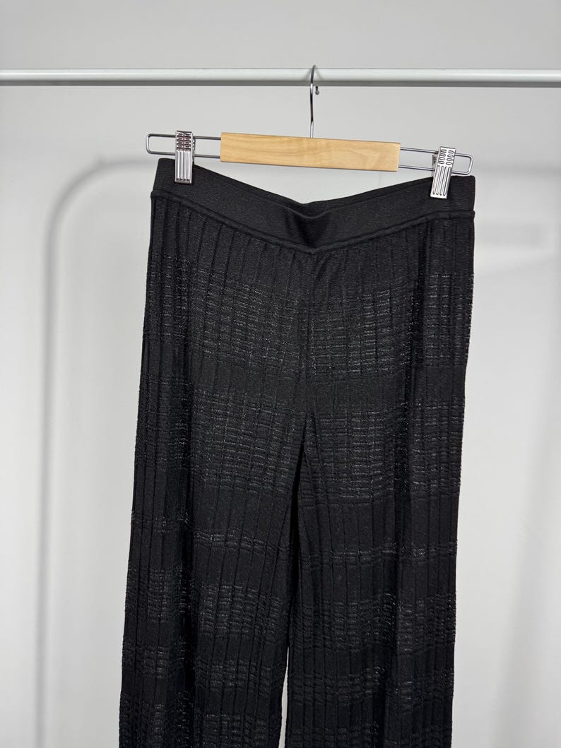 May include: Black, wide-leg pants with a textured, ribbed design. The pants are hanging on a wooden hanger with a silver metal clip. The fabric has a subtle sheen, and the pants appear to be made of a lightweight material.