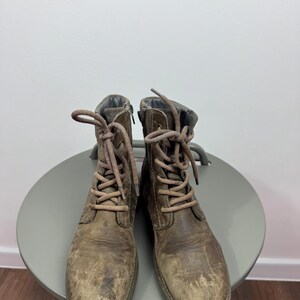 K&ouml;nnte beinhalten: Ein Paar abgenutzte braune Schn&uuml;rstiefel aus Leder mit runder Zehenpartie, mittelhohem Schaft und passenden braunen Schn&uuml;rsenkeln. Die Stiefel haben eine abgenutzte, strukturierte Oberfl&auml;che mit sichtbaren Kratzern und Farbvariationen.