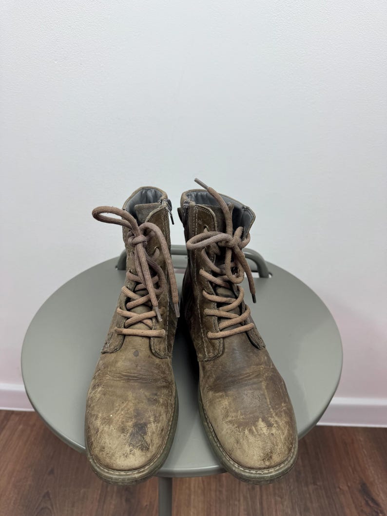 K&ouml;nnte beinhalten: Ein Paar abgenutzte braune Schn&uuml;rstiefel aus Leder mit runder Zehenpartie, mittelhohem Schaft und passenden braunen Schn&uuml;rsenkeln. Die Stiefel haben eine abgenutzte, strukturierte Oberfl&auml;che mit sichtbaren Kratzern und Farbvariationen.