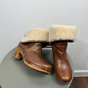 Könnte beinhalten: Braune Lederclogs mit einem Schafsfell-Manschetten. Die Clogs haben eine Holzsohle und einen Absatz mit Metallnieten. Die Stiefel werden auf einem grauen Tisch präsentiert. Die Schafsfell-Manschette ist hellbeige.