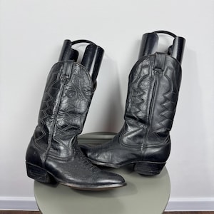 Puede incluir: Un par de botas de vaquero de cuero negro con costuras decorativas. Las botas se exhiben sobre un taburete gris. Las botas tienen una punta puntiaguda y un tacón apilado. Las botas son de estilo Western clásico.