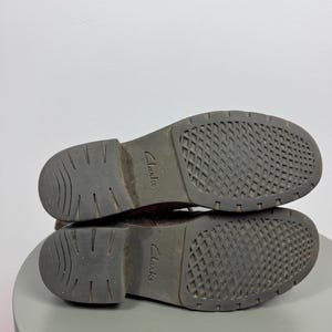 K&ouml;nnte beinhalten: Ein Paar dunkelbraune Stiefel in der Ansicht von unten, mit einer strukturierten Gummisohle, die ein Gittermuster im Vorderfu&szlig;bereich und geschwungene Rillen an der Ferse aufweist. Der Markenname Clarks ist mittig auf jeder Sohle eingepr&auml;gt.