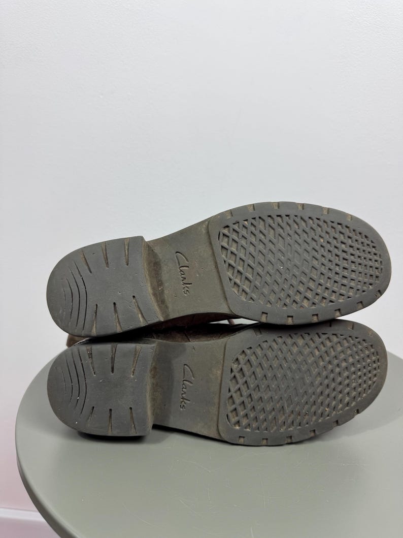 K&ouml;nnte beinhalten: Ein Paar dunkelbraune Stiefel in der Ansicht von unten, mit einer strukturierten Gummisohle, die ein Gittermuster im Vorderfu&szlig;bereich und geschwungene Rillen an der Ferse aufweist. Der Markenname Clarks ist mittig auf jeder Sohle eingepr&auml;gt.