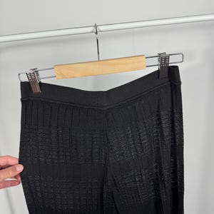 May include: Black knit pants with a textured, ribbed design, hanging on a wooden hanger. The pants have a high waist and a wide leg, with a subtle sheen. The fabric appears to be lightweight and suitable for warmer weather.