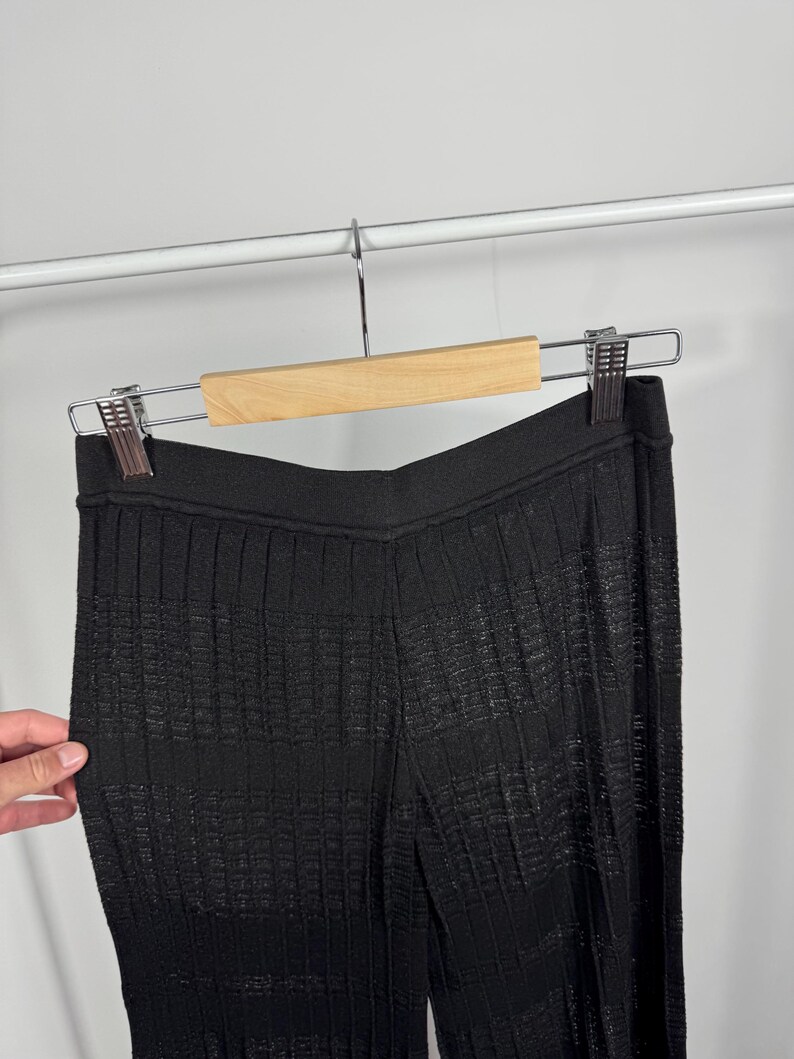 May include: Black knit pants with a textured, ribbed design, hanging on a wooden hanger. The pants have a high waist and a wide leg, with a subtle sheen. The fabric appears to be lightweight and suitable for warmer weather.