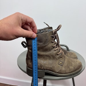 K&ouml;nnte beinhalten: Ein Paar braune Schn&uuml;rstiefeletten mit seitlichem Rei&szlig;verschluss und einer Zugschlaufe an der Ferse. Ein blaues Ma&szlig;band liegt an der Seite des Stiefels an und zeigt eine H&ouml;he von etwa 16,5 cm von der Sohle bis zum oberen Rand an.