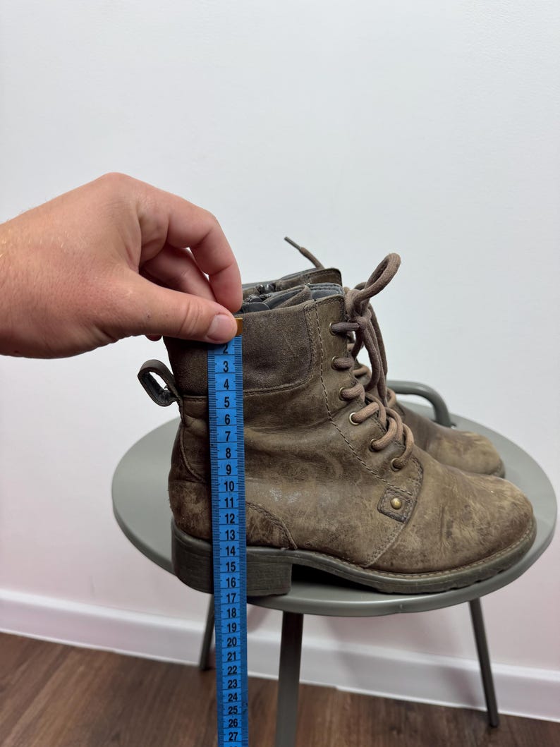 K&ouml;nnte beinhalten: Ein Paar braune Schn&uuml;rstiefeletten mit seitlichem Rei&szlig;verschluss und einer Zugschlaufe an der Ferse. Ein blaues Ma&szlig;band liegt an der Seite des Stiefels an und zeigt eine H&ouml;he von etwa 16,5 cm von der Sohle bis zum oberen Rand an.