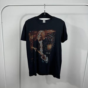 Puede incluir: Una camiseta negra de manga corta con un estampado gráfico de Kurt Cobain tocando la guitarra. El texto "YOU KNOW YOU'RE RIGHT" está impreso en letras blancas en el lado derecho del gráfico, con el nombre "COBAIN" impreso debajo.