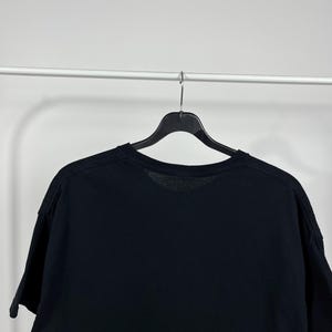 Peut inclure: Un t-shirt noir &agrave; manches courtes suspendu &agrave; un cintre noir sur fond blanc. Le t-shirt est fait d'une mati&egrave;re douce et a un col rond. L'image est un plan moyen, se concentrant sur la partie sup&eacute;rieure du t-shirt.