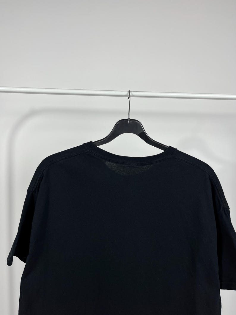 Peut inclure: Un t-shirt noir &agrave; manches courtes suspendu &agrave; un cintre noir sur fond blanc. Le t-shirt est fait d'une mati&egrave;re douce et a un col rond. L'image est un plan moyen, se concentrant sur la partie sup&eacute;rieure du t-shirt.