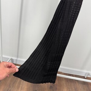 May include: A black, ribbed knit garment with a textured pattern. The fabric is folded, revealing the interior texture and construction. The garment is displayed against a white background and a wooden floor.
