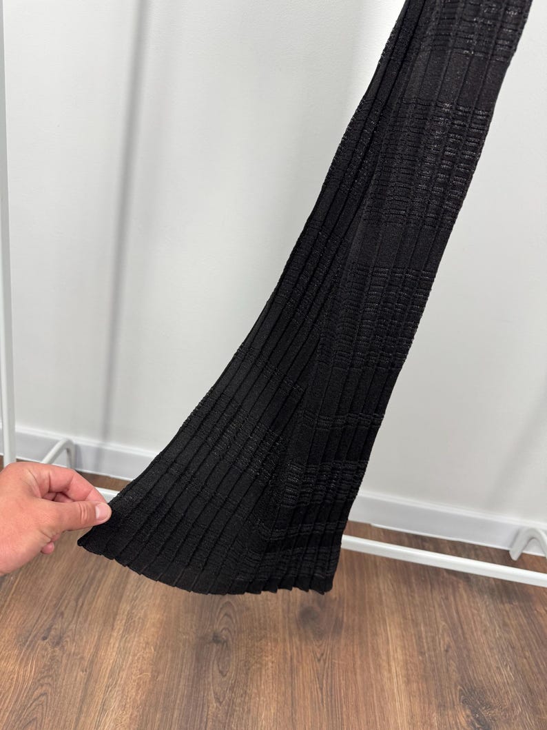 May include: A black, ribbed knit garment with a textured pattern. The fabric is folded, revealing the interior texture and construction. The garment is displayed against a white background and a wooden floor.