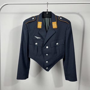 Puede incluir: Chaqueta militar azul marino oscuro con botones plateados e insignias en los hombros. La chaqueta tiene un diseño recortado y presenta dos bolsillos delanteros y un emblema plateado en el pecho. Los detalles del cuello y los hombros tienen detalles dorados.