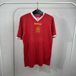 May include: A red short-sleeved jersey with a white V-neck collar and white sleeve cuffs. The shirt features the yellow Reebok logo, a yellow Liverpool FC crest with four stars above it, and the word Carlsberg embossed on the front chest area.