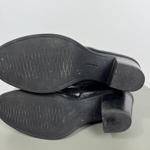 Pu&ograve; includere: Stivaletti in pelle nera con tacco a blocco. Le suole sono strutturate con un motivo a diamante. Il marchio "Clarks" &egrave; visibile sulla suola. Gli stivali sono su una superficie grigio chiaro.
