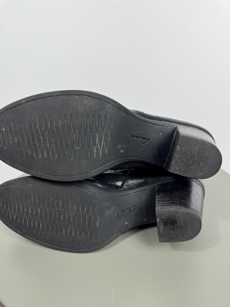 Pu&ograve; includere: Stivaletti in pelle nera con tacco a blocco. Le suole sono strutturate con un motivo a diamante. Il marchio "Clarks" &egrave; visibile sulla suola. Gli stivali sono su una superficie grigio chiaro.