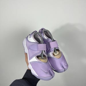 May include: Pair of lavender and white athletic shoes with a split-toe design. The shoes feature a hook-and-loop strap, a white sole, and a small Nike swoosh logo.