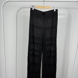 May include: Black, wide-leg pants with a textured, open weave design. The pants are hanging on a wooden hanger suspended from a white metal clothing rack. The fabric appears to be lightweight and suitable for warmer weather.
